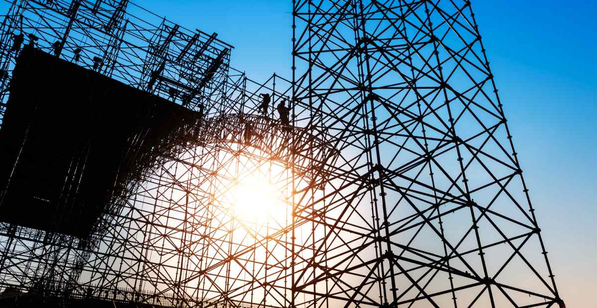 ● Workers on construction scaffolding, seen from below with a blue sky and sun flare shining through.